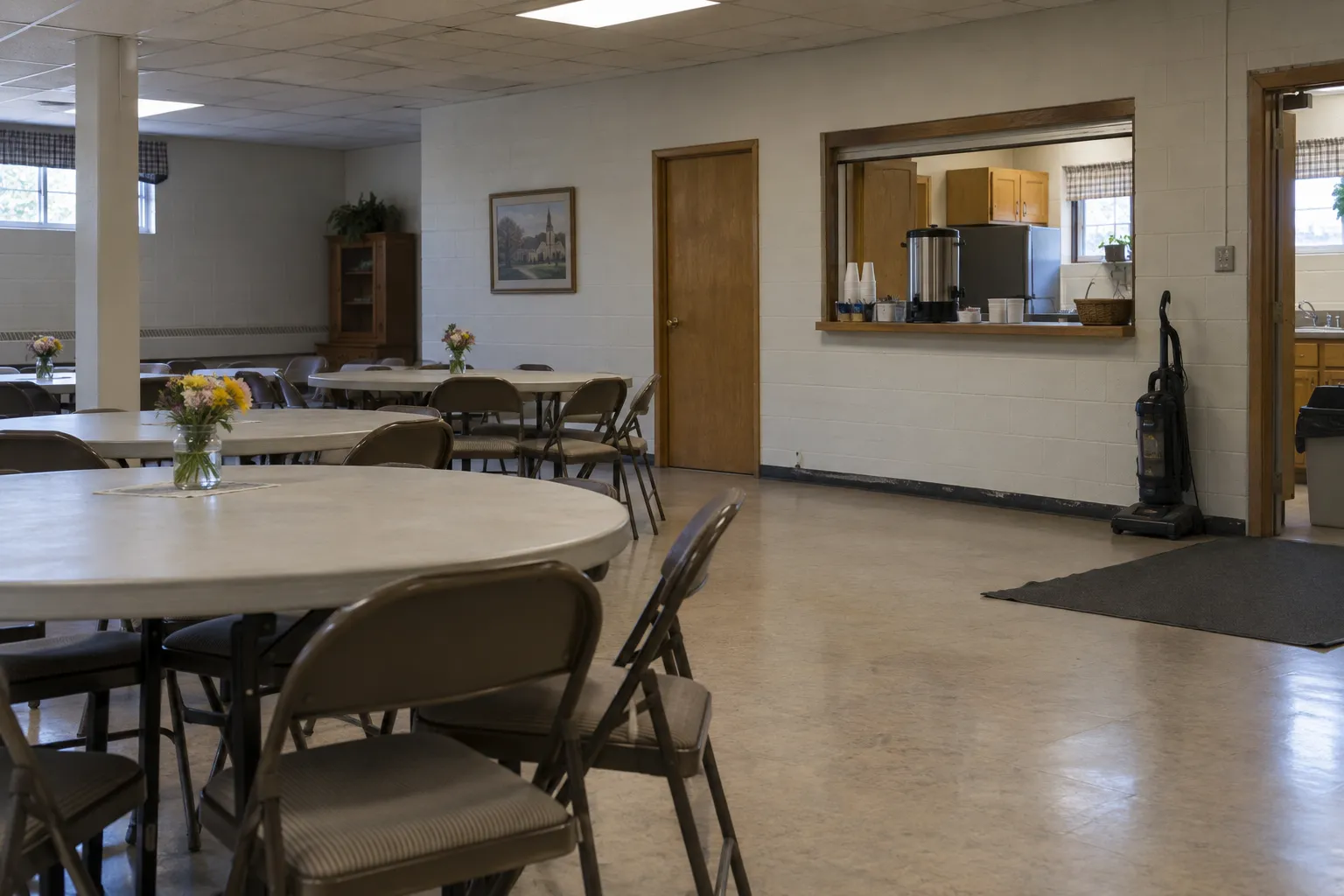 A church fellowship hall with round tables and a vacuum by the kitchen pass-through