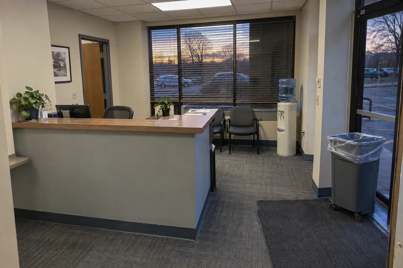 Reception area of a small suburban office suite at dusk after cleaning, with vacuumed carpet tiles, a wiped counter, a water cooler and a fresh trash liner