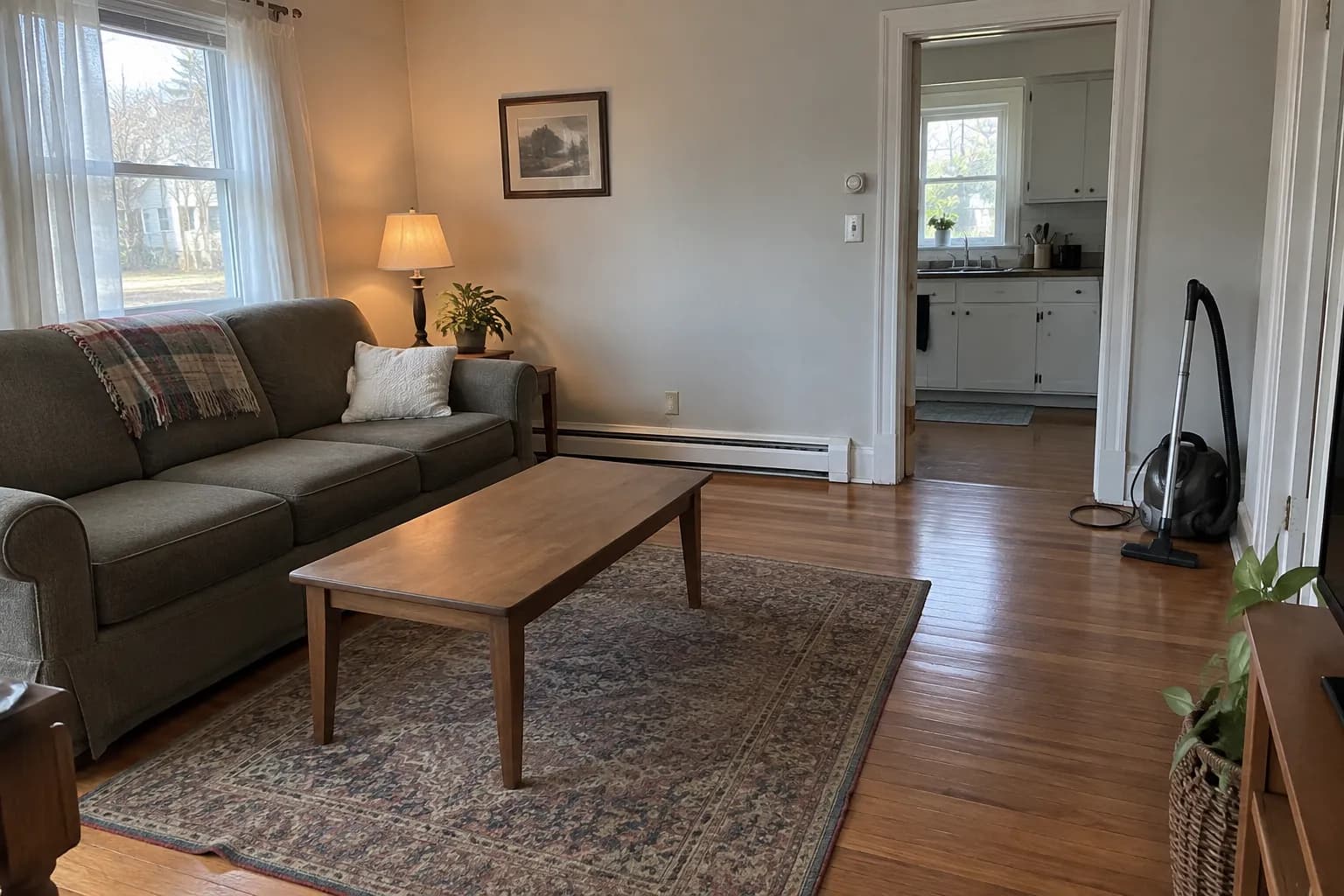 Tidy living room in a modest older house with polished hardwood floors, a patterned rug and a canister vacuum by the kitchen doorway