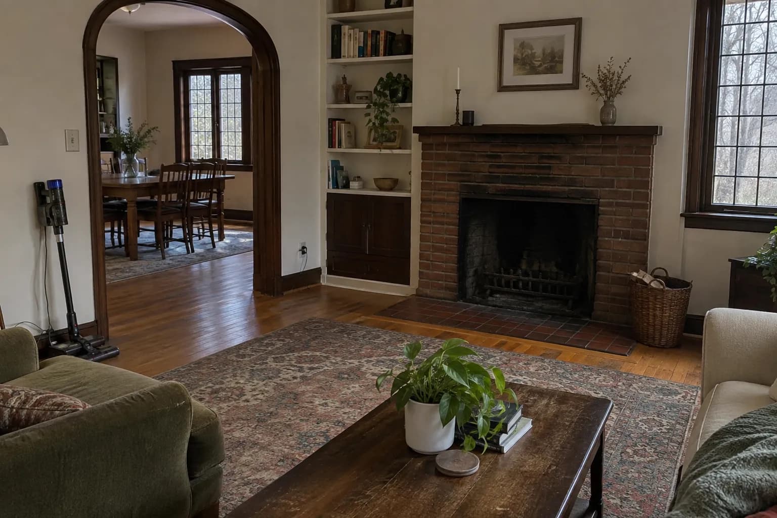 Living room of a 1920s Tudor-style house after cleaning, with leaded-glass windows, a fireplace, built-in shelves, an arched doorway and a stick vacuum