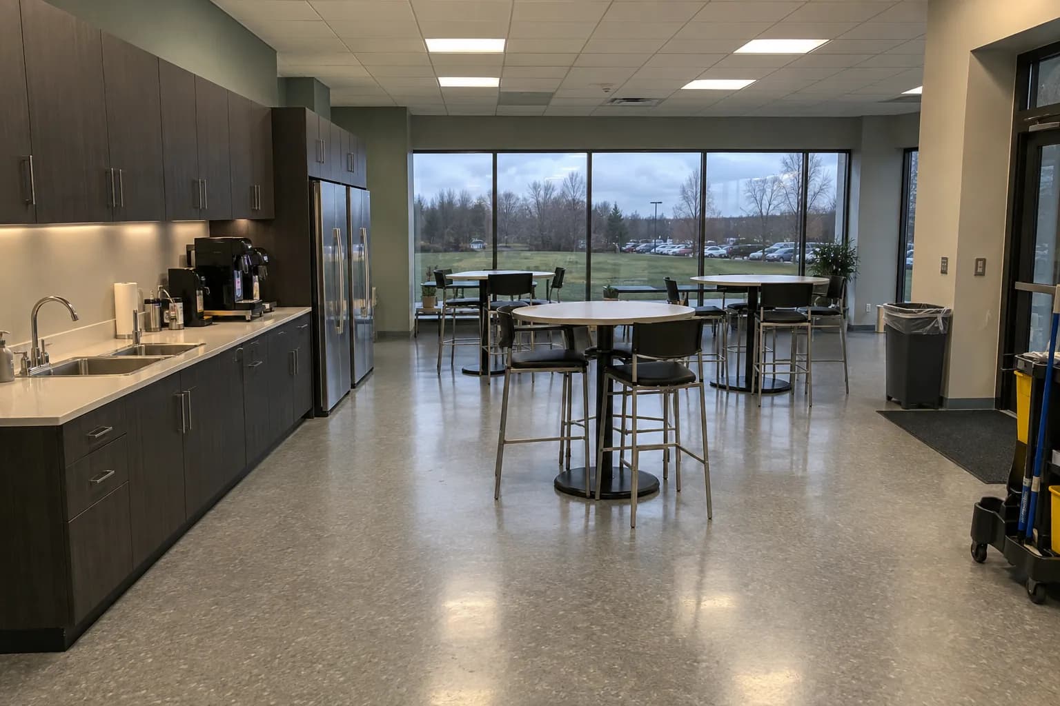 Staff kitchen and cafe area in a modern office building after an evening cleaning, with a long counter and sinks, stainless refrigerators, high-top tables with stools, a freshly mopped floor, windows onto a lawn and parking lot, and a janitorial cart by the entrance