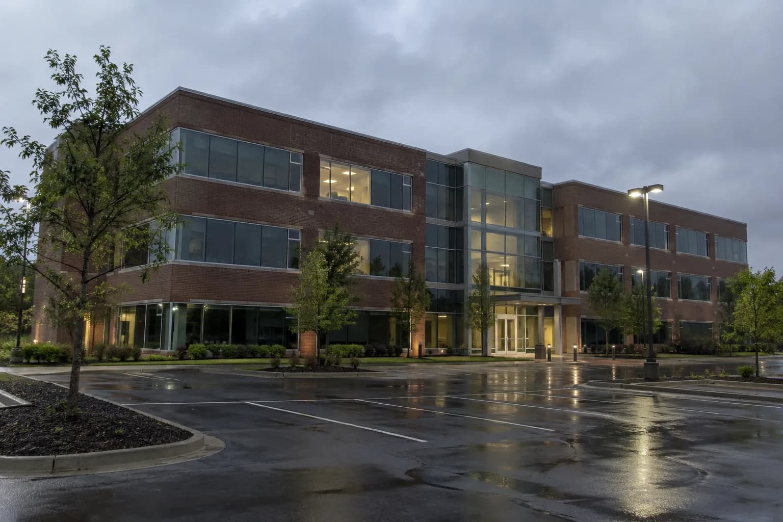 A brick-and-glass suburban office building at dusk across a rain-wet parking lot