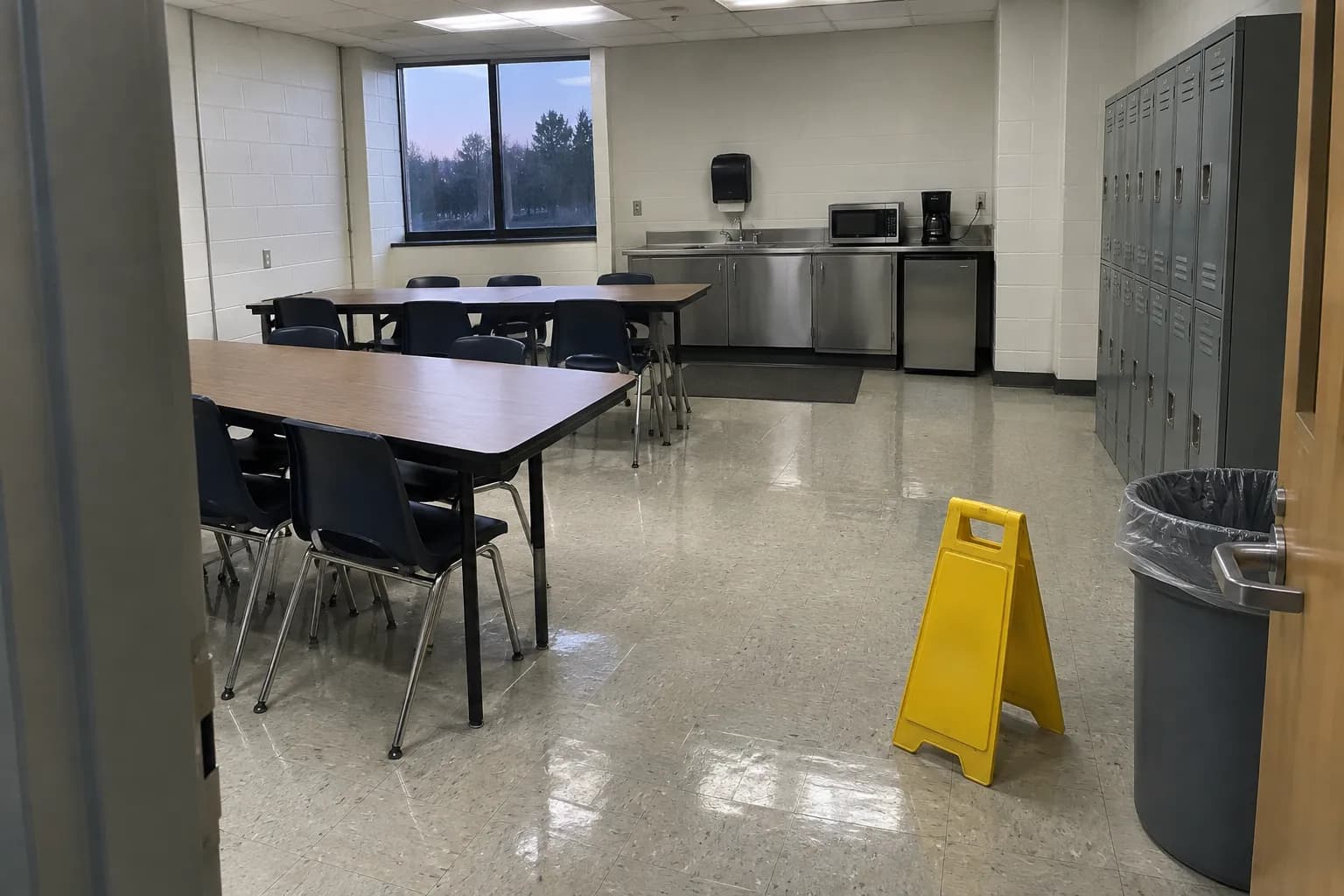 Employee break room in a light-industrial building after an evening cleaning, with wiped tables, a stainless counter with a microwave, gray lockers, a mopped tile floor and a yellow wet-floor sign