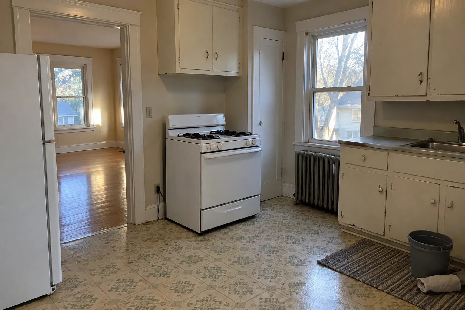 Kitchen of an empty older apartment after a move-out cleaning, with painted cabinets, a white range and refrigerator, a radiator under the window and a patterned vinyl floor