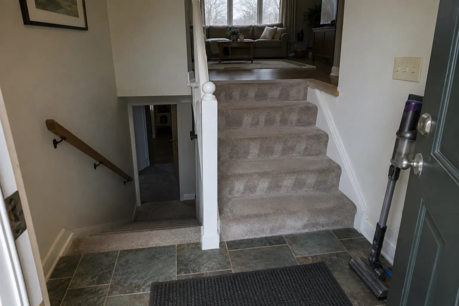 Split-level entry of a raised ranch house after a routine cleaning, with a tiled landing and door mat, freshly vacuumed carpeted stairs leading up to the living room and down to the lower level, and a cordless vacuum by the door