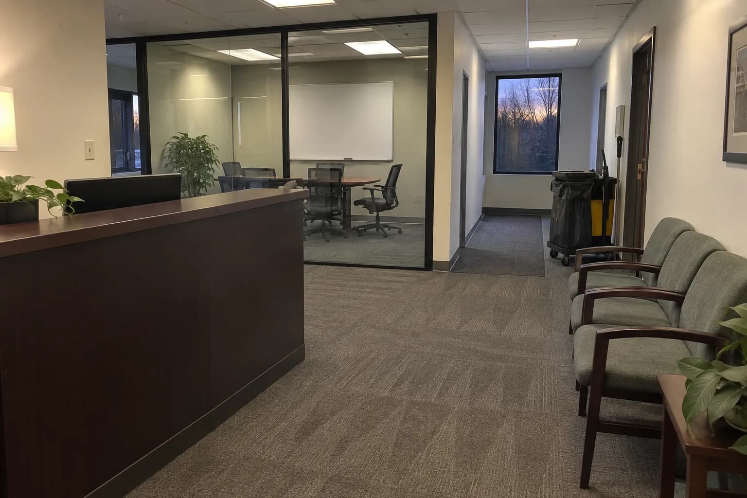 Reception area of a small professional office at dusk after cleaning, with vacuumed carpet, waiting chairs, a glass-walled conference room and a janitorial cart