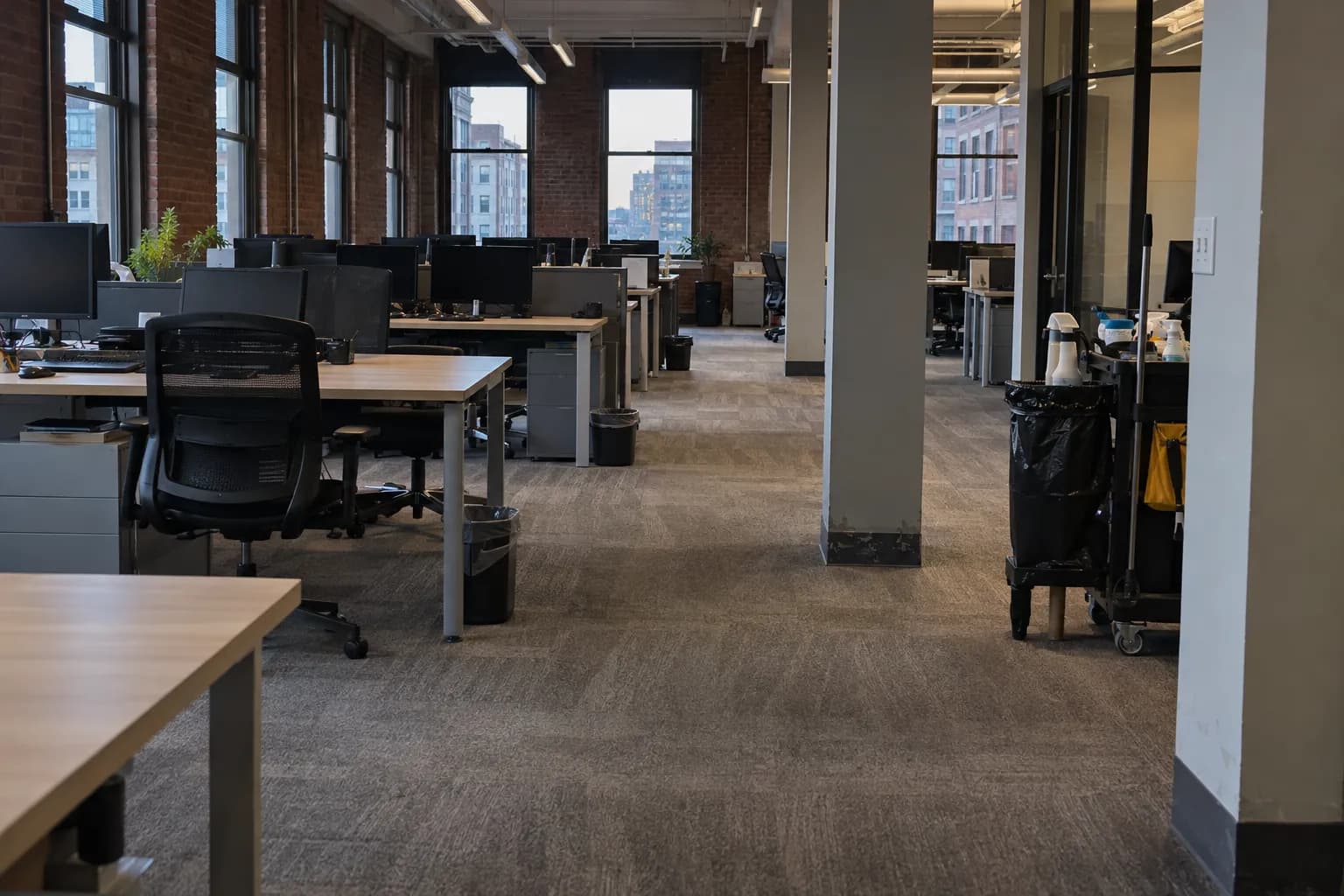 Open-plan office in an older brick building after an evening cleaning, with vacuumed carpet and a janitorial cart by a pillar