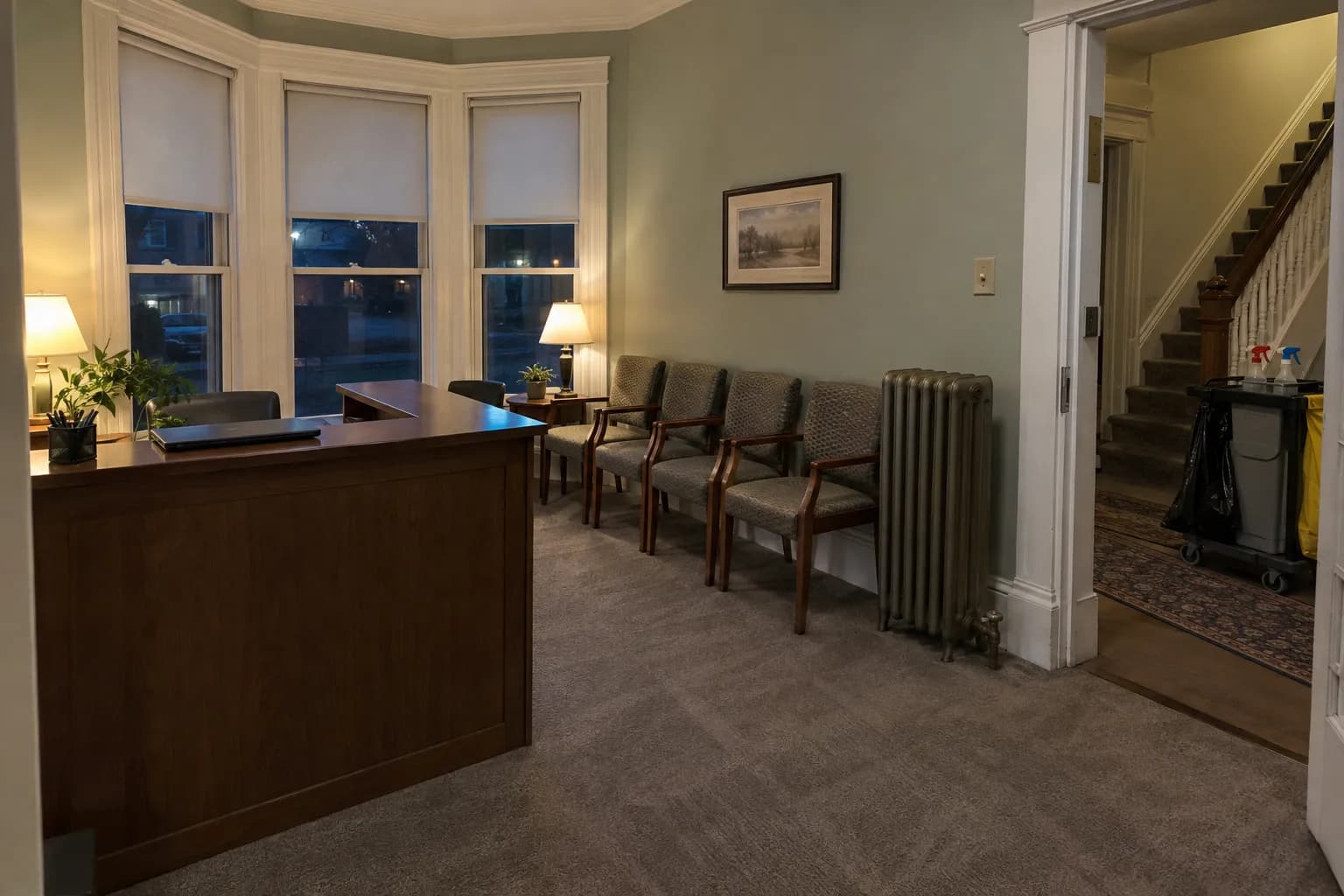 Reception room of a small office in a converted Victorian house after an evening cleaning, with a bay window, a wooden reception desk, a row of waiting chairs, a cast-iron radiator, freshly vacuumed carpet and a janitorial cart in the hall by the stairs