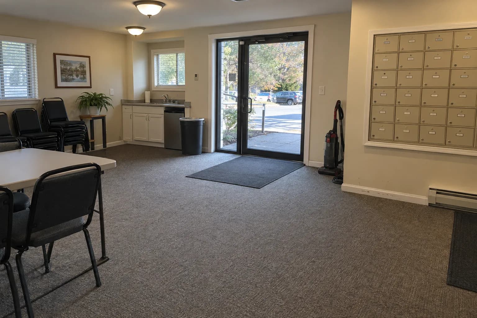 Community room of a condominium complex after cleaning, with carpet, a folding table and stacked chairs, a small kitchenette, a wall of cluster mailboxes and an upright vacuum by the glass door