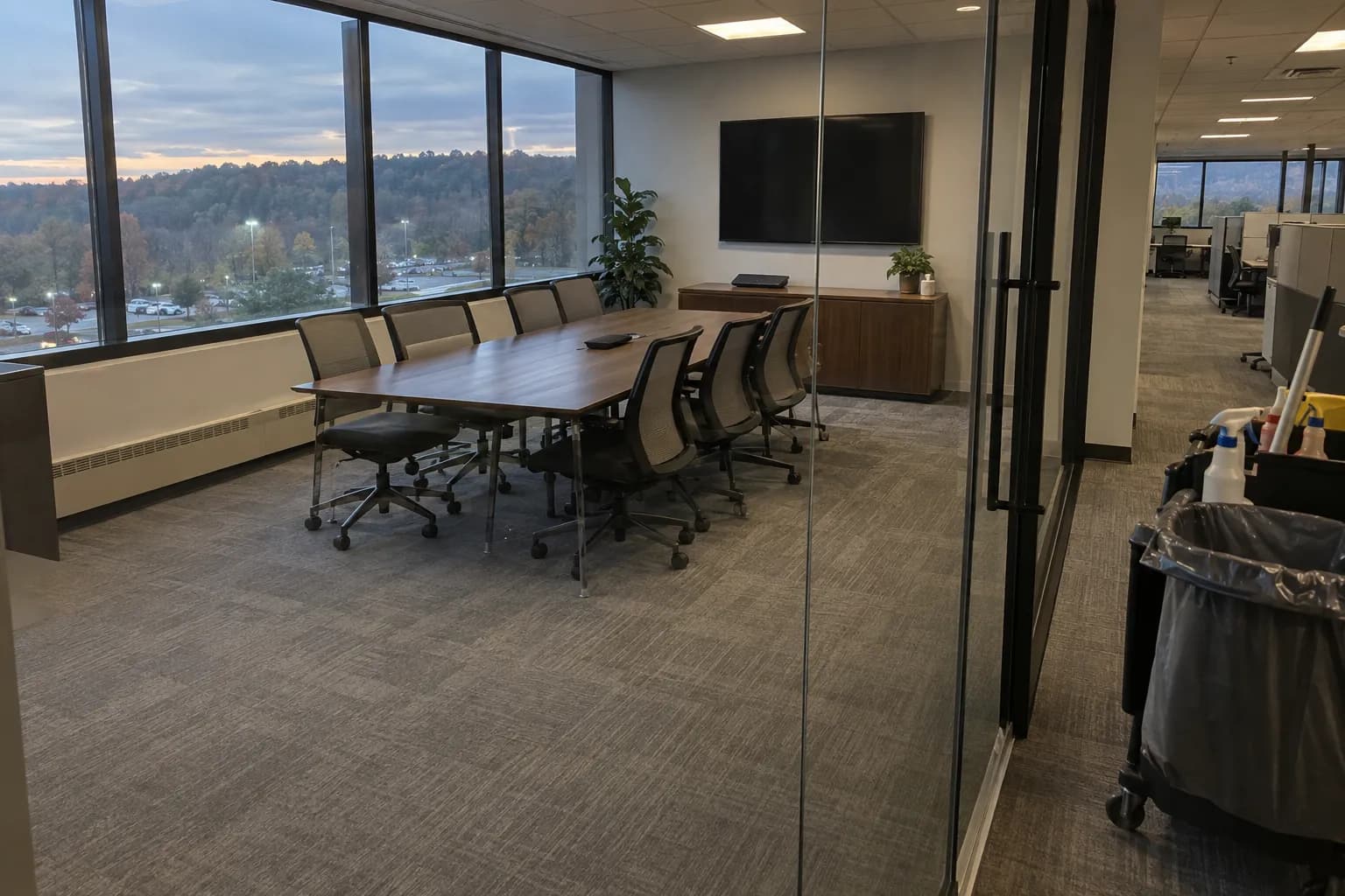 Conference room on an upper floor of a suburban office building after an evening cleaning, with mesh chairs pushed in around a long table, freshly vacuumed carpet, a wooded hillside and lit parking lot beyond the windows, and a janitorial cart in the corridor