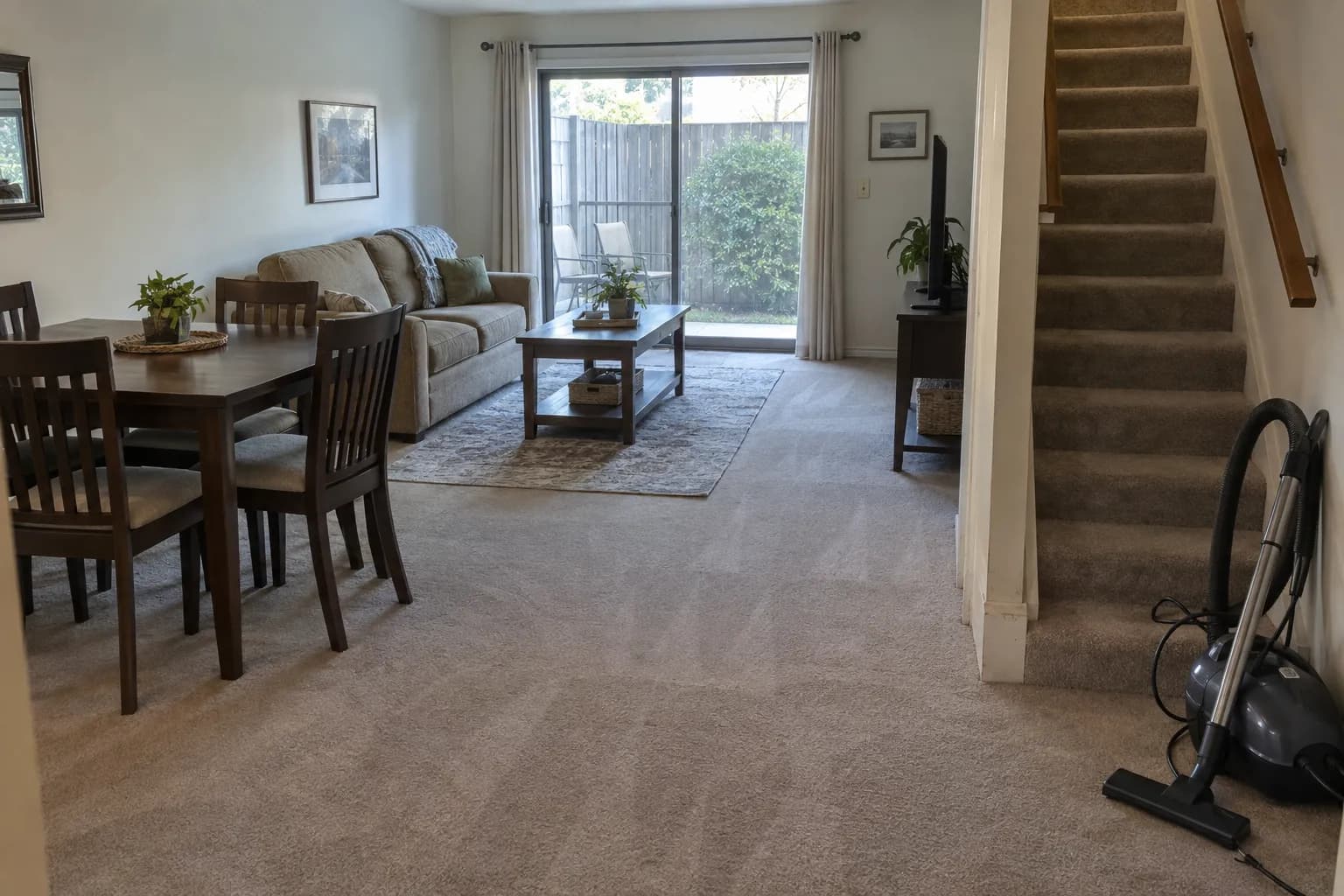 Living and dining room of a two-story townhouse after cleaning, with vacuum lines in beige carpet, a carpeted staircase, a sliding door to a fenced patio and a canister vacuum at the foot of the stairs