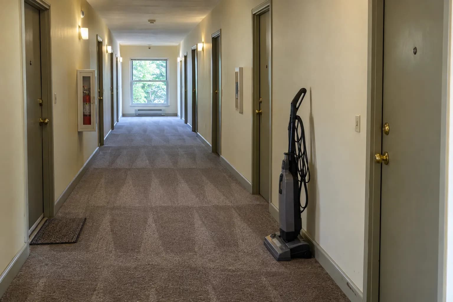 Carpeted interior hallway of a garden-style apartment building with fresh vacuum lines, a row of unit doors and an upright vacuum against the wall