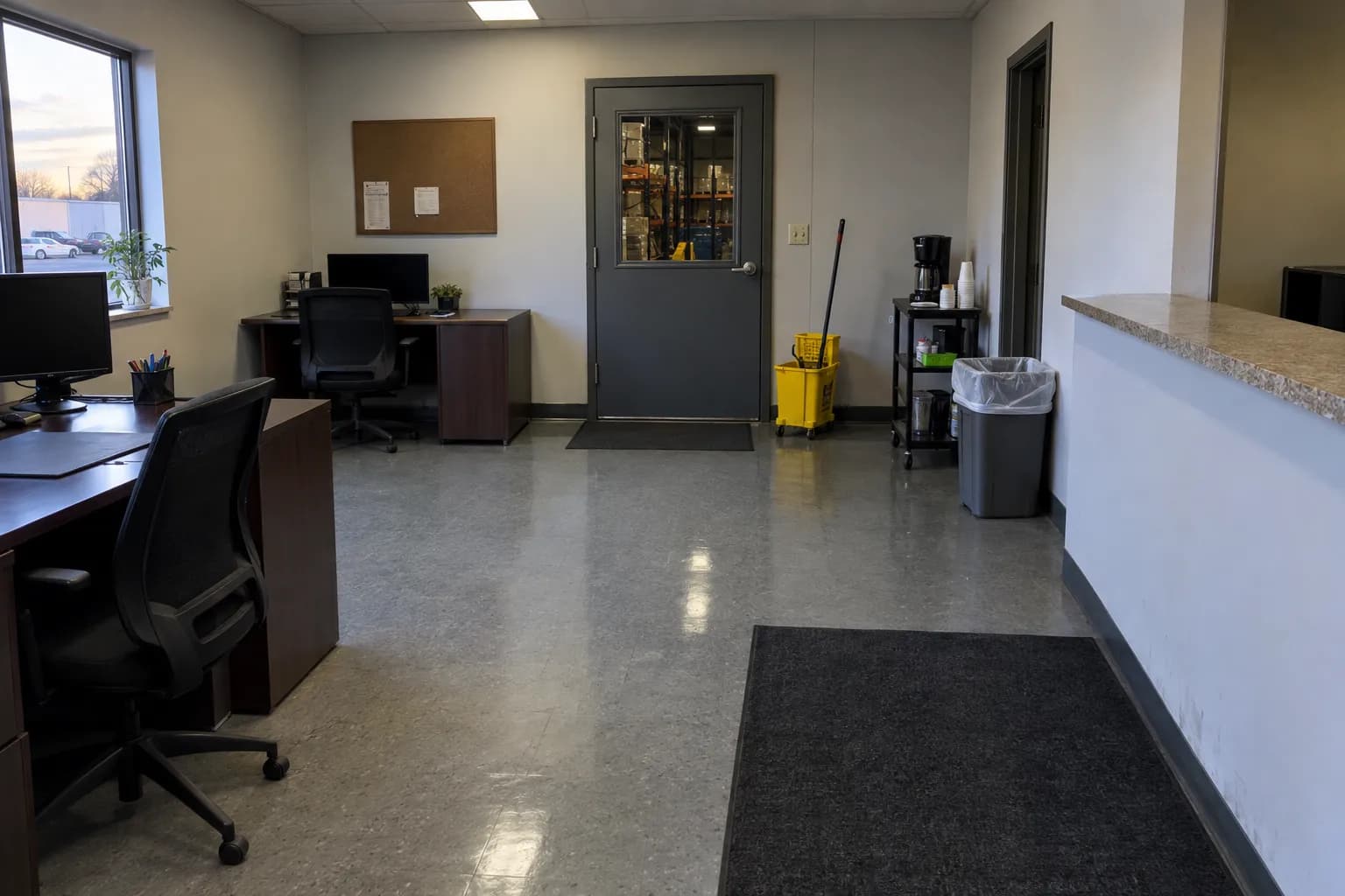 Front office of a small light-industrial business after an evening cleaning, with two desks, a service counter, a freshly mopped tile floor, a coffee cart, a trash can with a fresh liner, a yellow mop bucket and a door looking into a warehouse